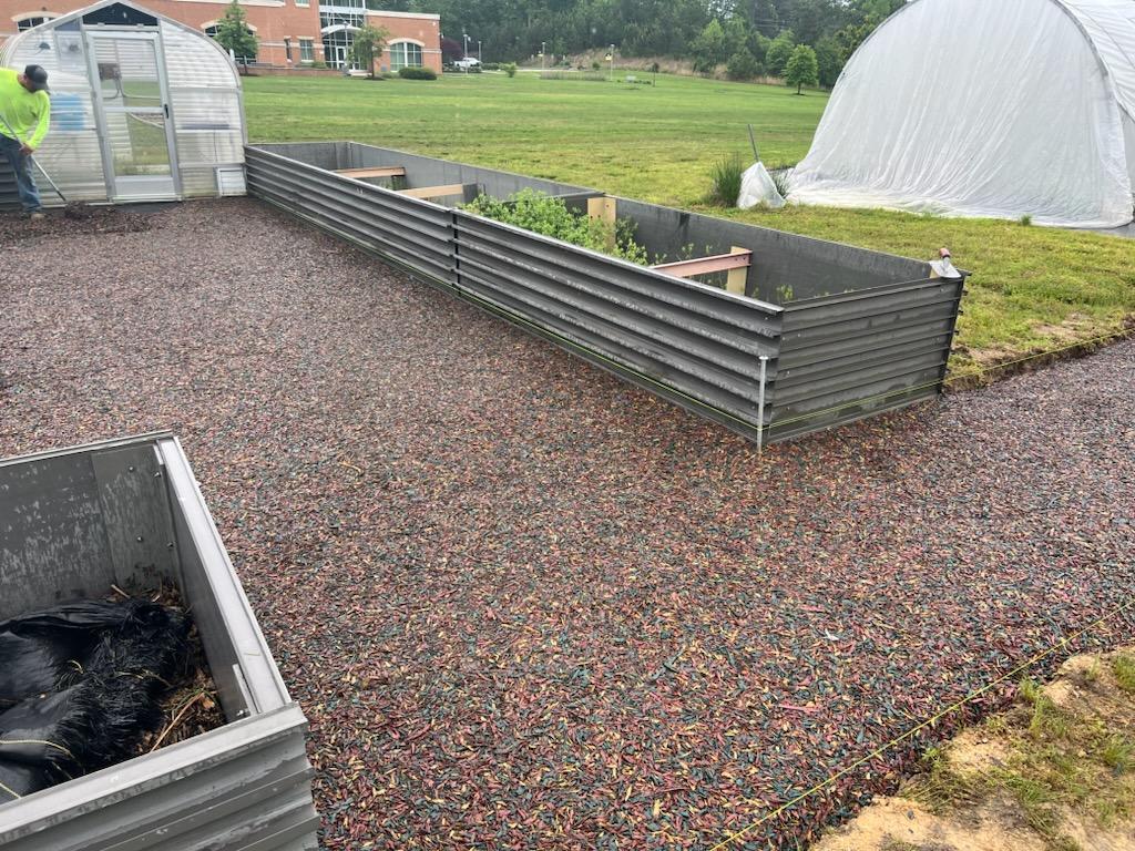 Garden beds made of metal with a gravel pathway, set in a green outdoor area near a greenhouse.