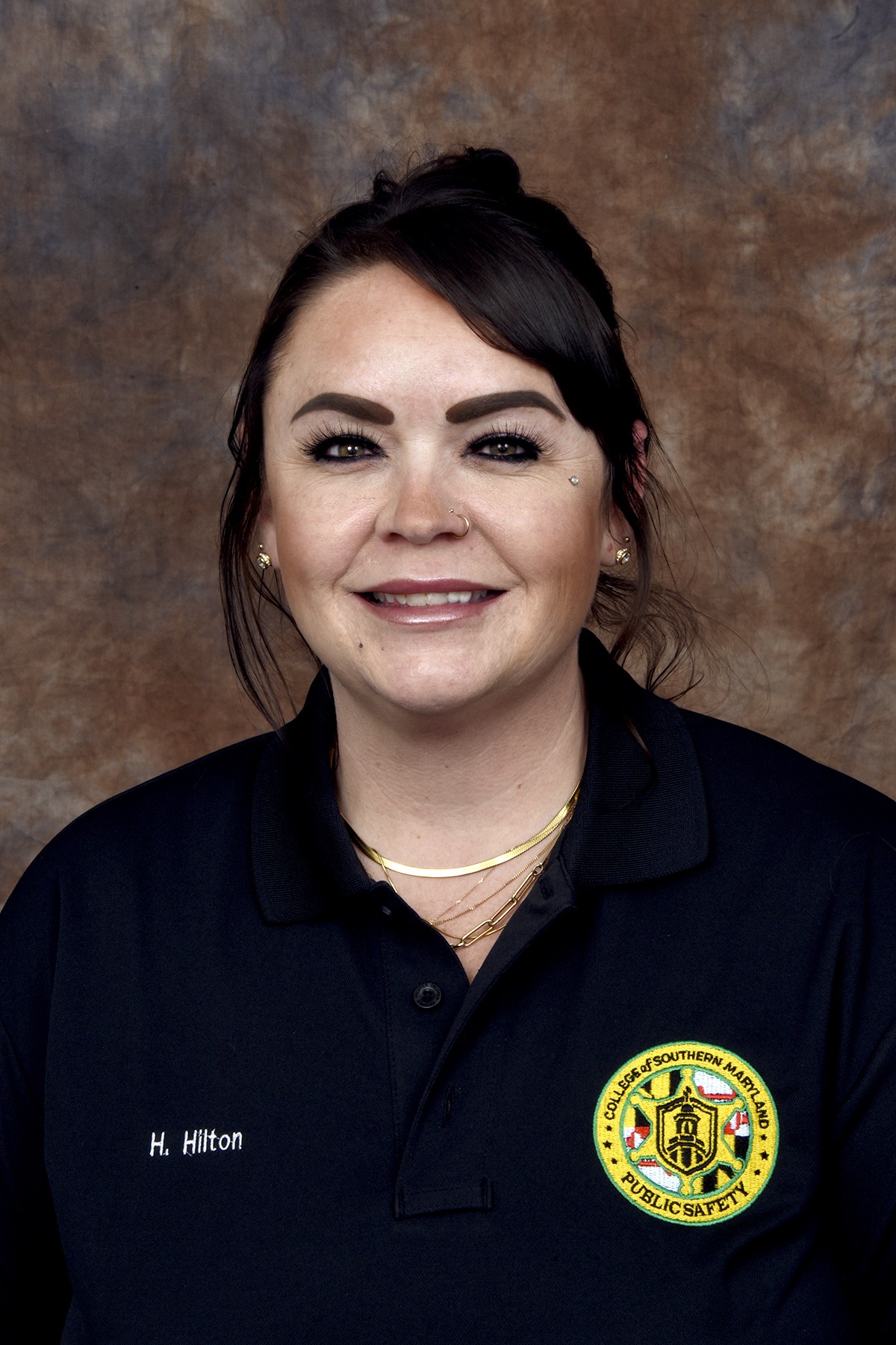 A portrait of a woman in a uniform shirt with a public safety logo, wearing necklaces and a purple ribbon pin.