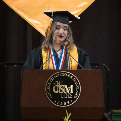 A young blonde woman in graduation regalia stands behind a wooden podium with the seal of the college