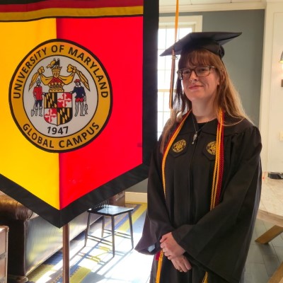 Danielle Barber from CSM's Class of 2022 stands in graduation regalia next to a banner with the crest of University of Maryland Global Campus