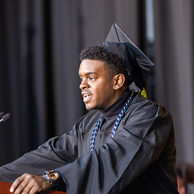 CSM graduate Kaleb Bennett stands in profile, speaking at the podium during commencement and wearing a graduation cap and gown. 