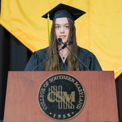 CSM graduate Kara Novak stands at a wooden podium emblazoned with the college's crest, wearing graduation regalia, and looking slightly to her left as she addresses the audience during her speech