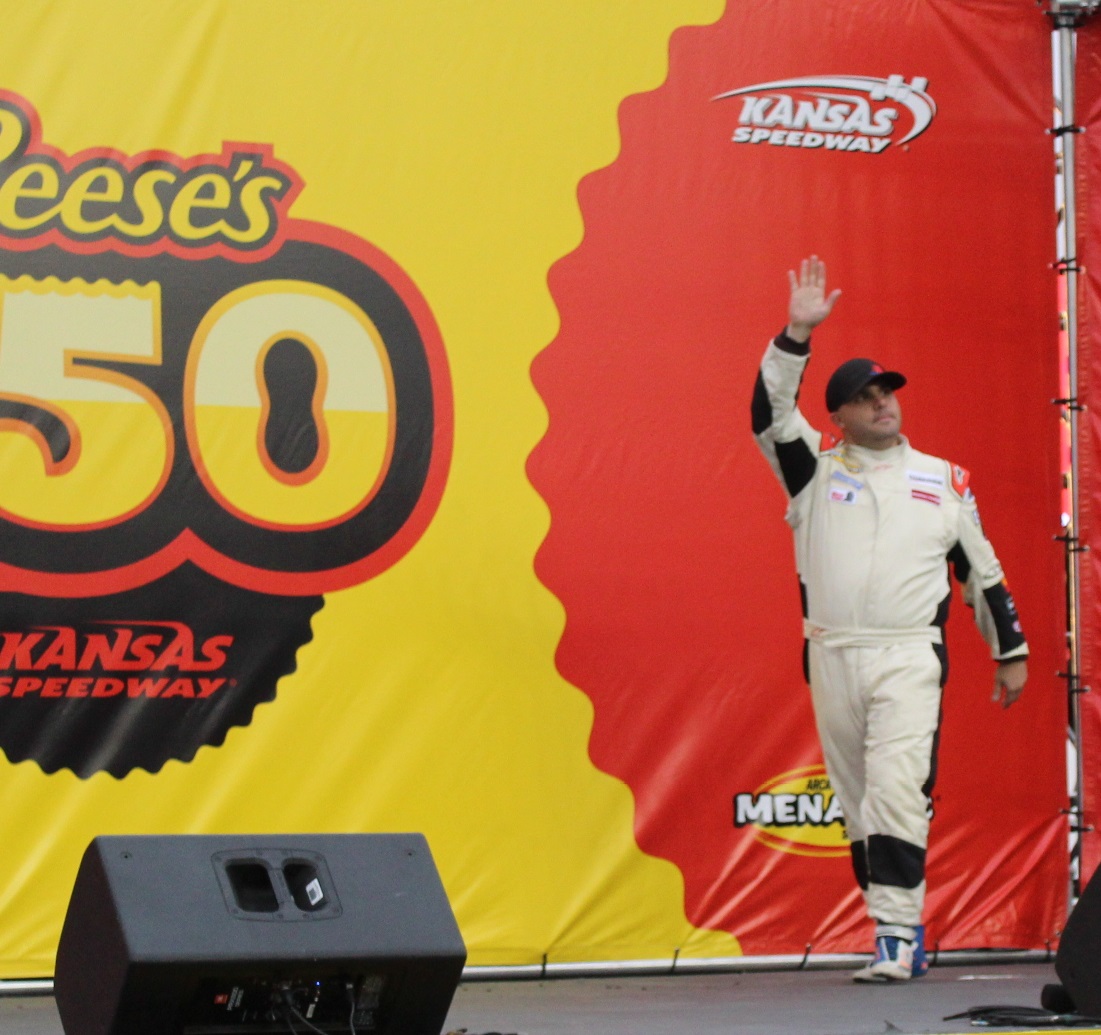 CSM graduate Kyle Lockrow, dressed in a racing jumpsuit, striding confidently onto a stage and waving, in front of a backdrop reading "Reese's 150 at Kansas Speedway"