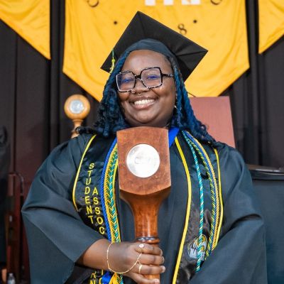 CSM graduate Markala Pharr stands in front of the podium and CSM banner at commencement, dressed in graduation cap and gown