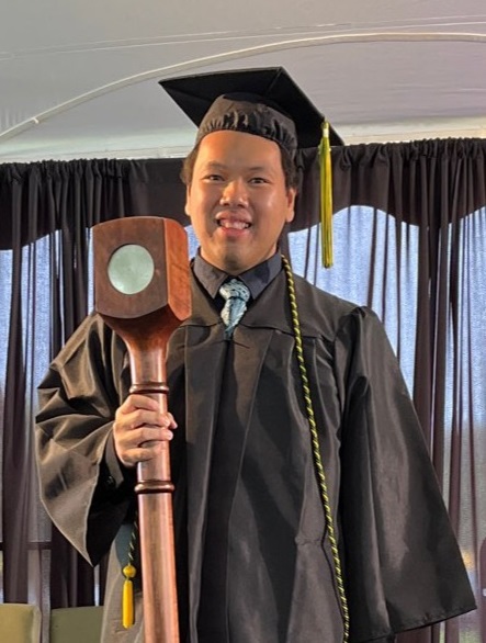 CSM alum Michael Chiong in graduation regalia and holding the college's ceremonial mace