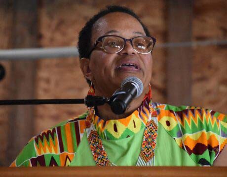 A woman wearing a vibrant traditional African outfit and large earrings speaks at a microphone.