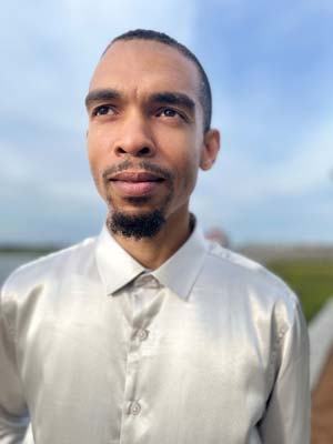 A man in a silver shirt standing outdoors with a thoughtful expression.