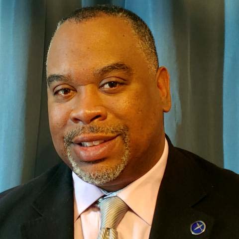 A professional headshot of a man wearing a suit and tie, smiling confidently against a blue backdrop.