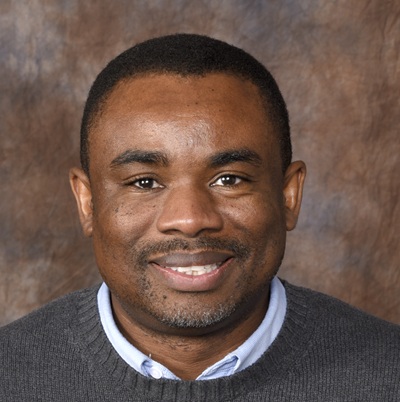 Assistant Professor Amos Dike smiles at the camera. He's wearing a dark blue sweater over a light blue collared shirt.