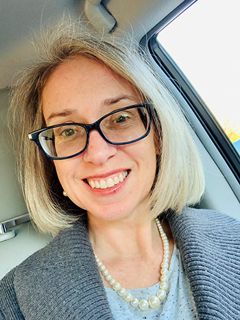 Smiling woman with blonde hair and glasses taking a selfie inside a car, wearing a gray cardigan and pearl necklace.
