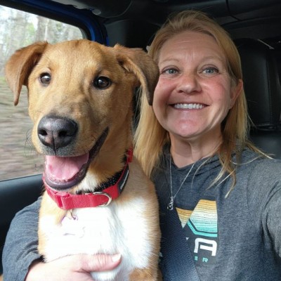 A selfie of CSM professor Dr. April Rollins, a blonde woman who is smiling at the camera, wearing a gray sweatshirt, and with her arm around a dog sitting in her lap looking cute and happy.