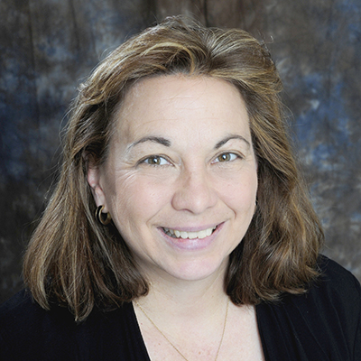 A professional headshot of Laurie Cangelosi. She is sitting in front of a blue and gray backdrop, smiling and wearing a black top.