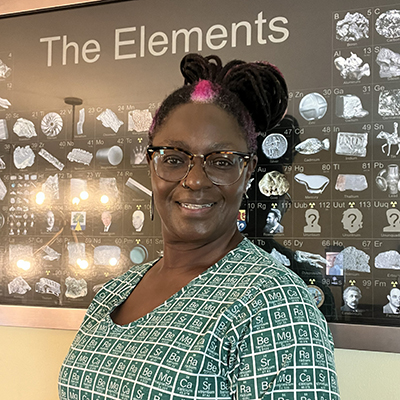 Chemistry professor Valerie Shelton smiles at the camera. She is standing in front of a wall poster with the periodic table of elements and wearing a green dress printed with the periodic table.