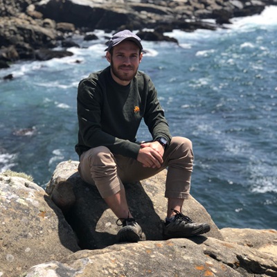 CSM professor Dr. Brandon Wilfong sits on a rock with a large body of water in the background, wearing a baseball cap, dark sweatshirt, and khaki pants.
