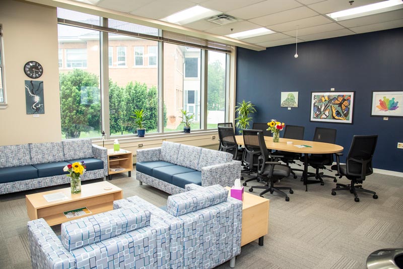 A modern, welcoming lounge area featuring blue sofas, a wooden table, and a colorful display of artwork on the walls. Natural light fills the space from large windows, complemented by indoor plants.