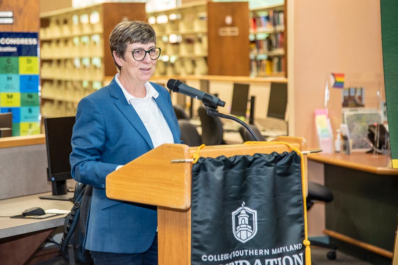A woman with glasses speaks at a podium with a banner for the College of Southern Maryland Foundation, set in a library.
