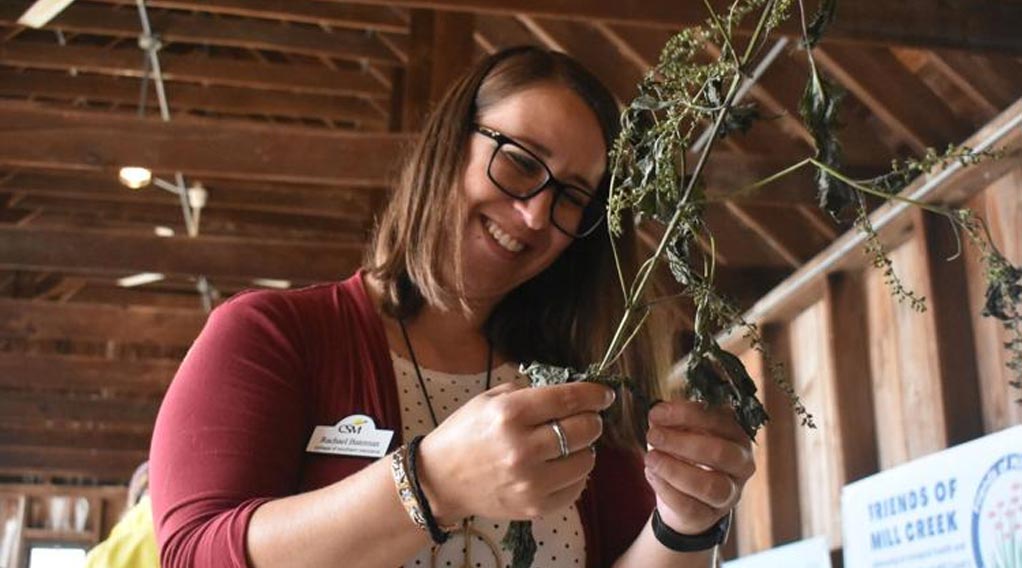 A woman with glasses smiles while inspecting dried ivy vines at a community event.