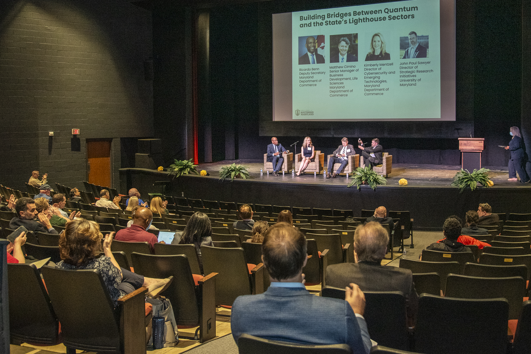 Panelists sit on stage with attendees listening in chairs