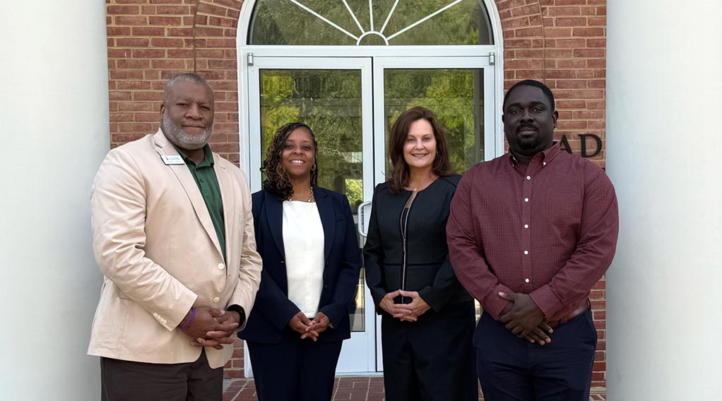 A group of four professionals poses for a photo outside a building, showcasing a diverse representation. They are standing in front of large glass doors with a welcoming demeanor.