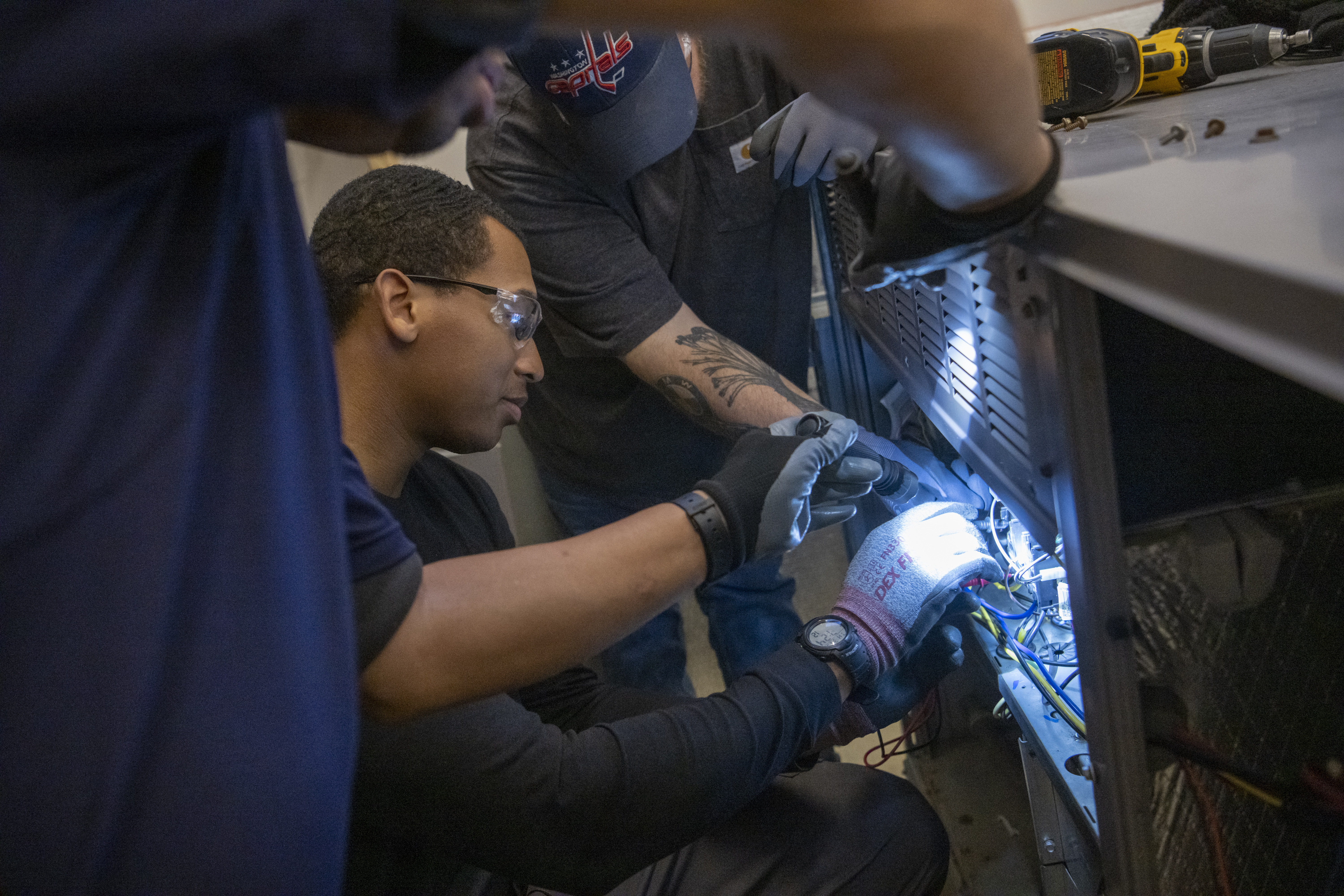 Three people work on HVAC system holding a flashlight to wires