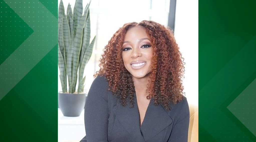 A smiling woman with curly brown hair, wearing a black outfit, seated beside a tall green plant in a bright indoor setting.