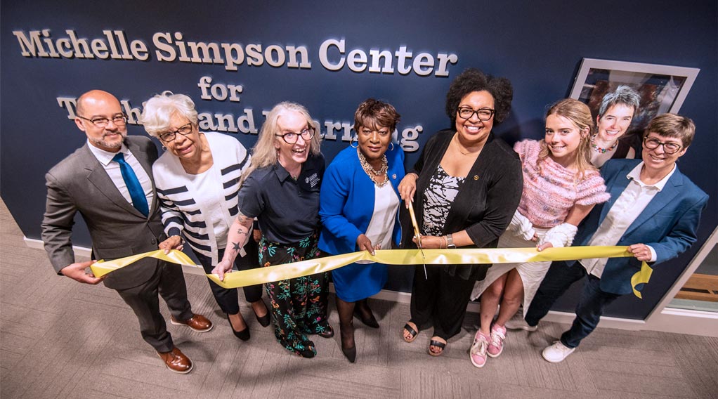 Eight people, four appearing to be women and four appearing to be men, smile while holding a yellow ribbon during a ribbon-cutting ceremony for the Michelle Simpson Center for Teaching and Learning. A woman in a blue blazer is cutting the ribbon.