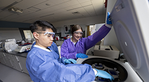 Two lab technicians wearing safety glasses and gloves work together at an open centrifuge in a laboratory.