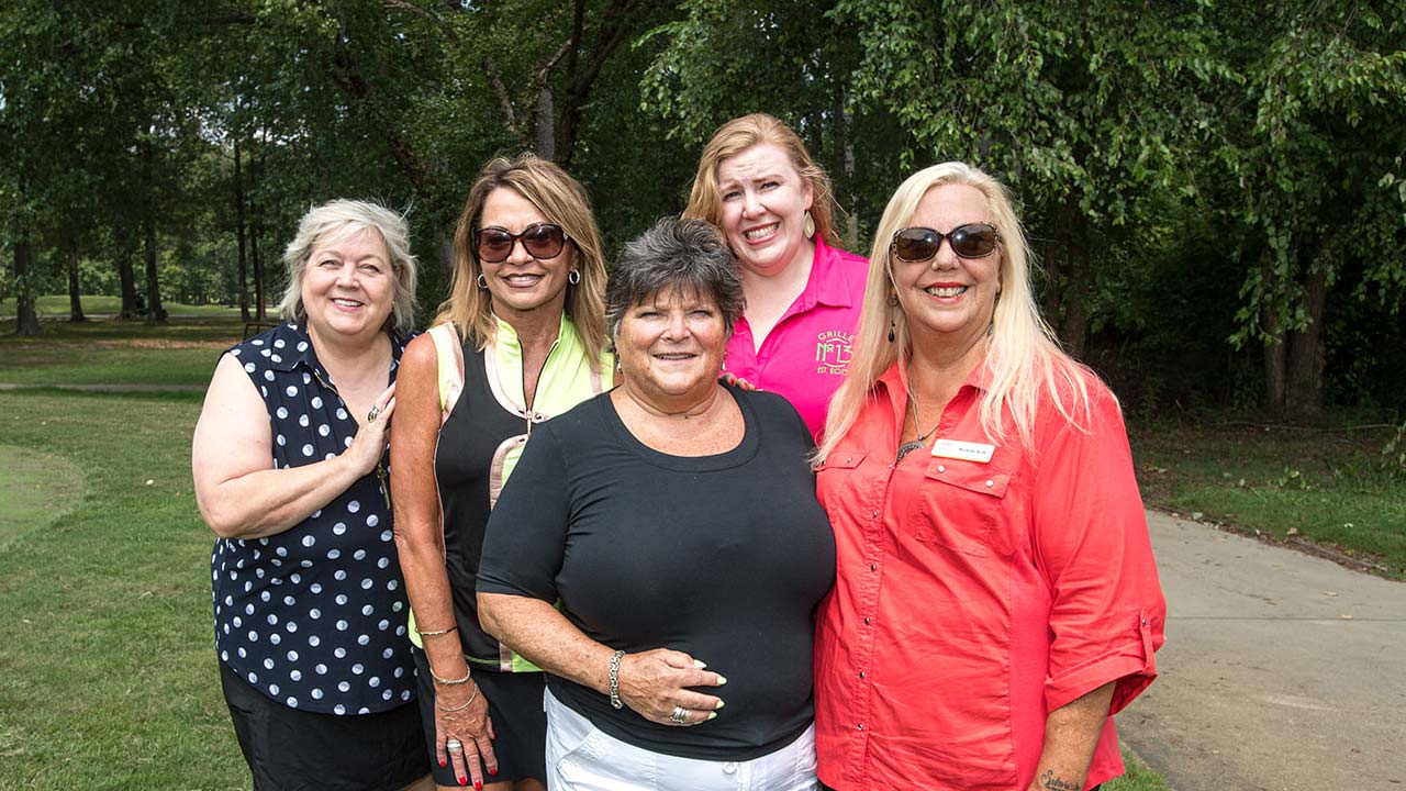 A group of five women smiling and posing together outdoors, dressed in casual attire.