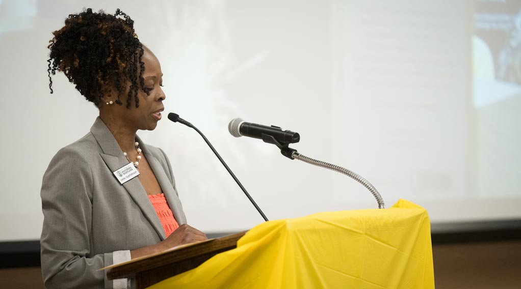 A woman speaks at a podium covered with a yellow cloth, with a microphone in front of her and a projection screen in the background.