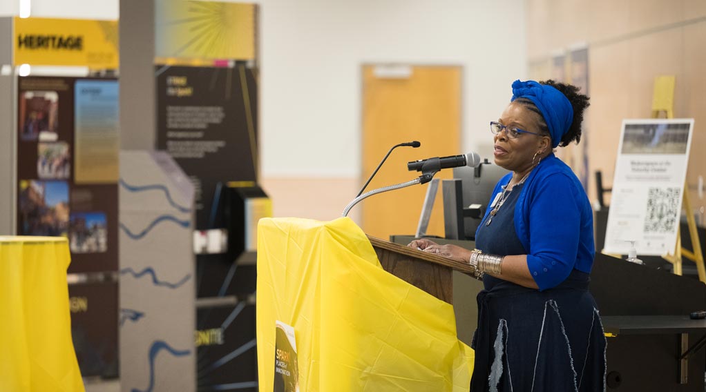 A woman in a blue outfit and headwrap speaks at a podium during an event, with display boards in the background.