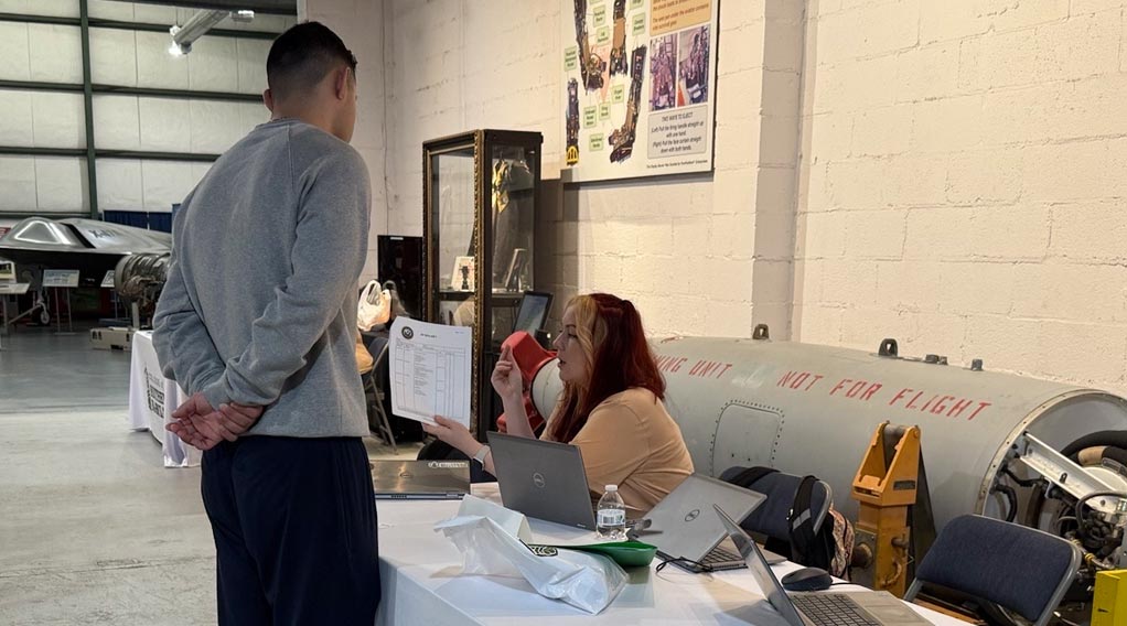 A woman sits at a table providing information to a man standing in front of her at a public event.