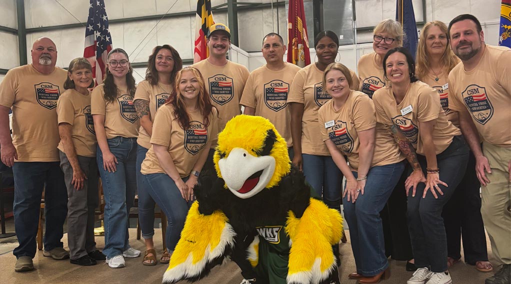 Group of staff and faculty in matching brown t-shirts reading 'serving those who serve' posing with the CSM Hawk mascot