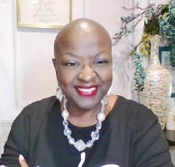 A smiling woman with a shaved head wearing a black shirt and a statement necklace, standing in an elegantly decorated room.